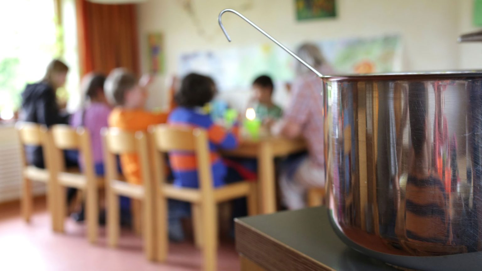 Kochtopf im Vordergrund, Kinder sitzen gemeinsam am Tisch im Hort.